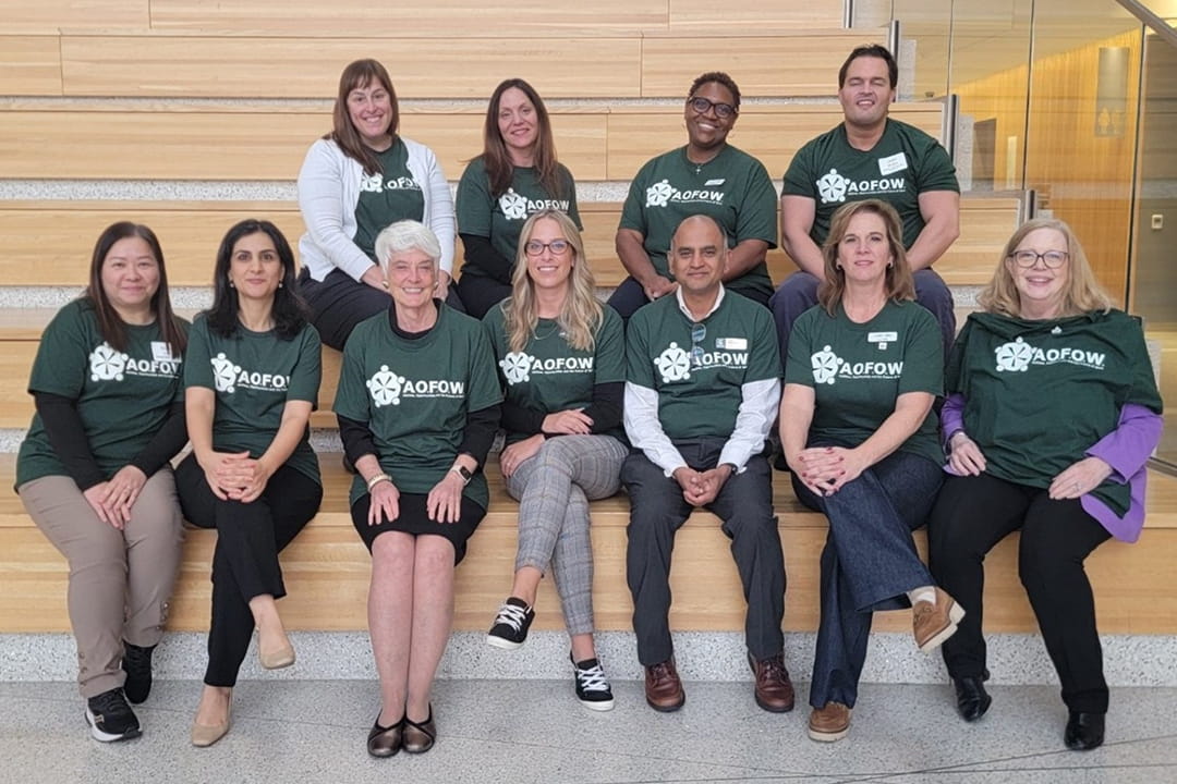 Group photo of the Abilities, Opportunities and Future of Work (AOFOW) conference team — eleven adults seated on indoor tiered wooden seating, wearing matching dark green AOFOW T-shirts and smiling at the camera.