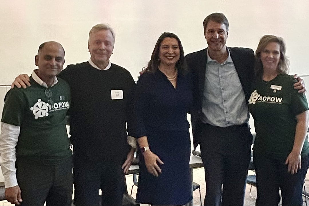 Five panelists standing together at the AOFOW conference — two wearing green AOFOW T-shirts and three in business attire — smiling with arms around each other after speaking on AI and technology.