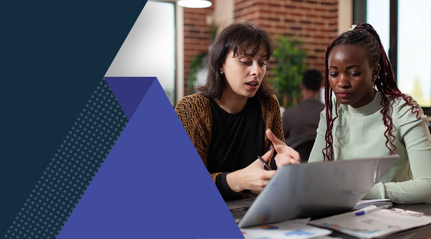 Two individuals sit at a desk collaborating over a laptop, with documents and office supplies spread out on the table. A brick interior wall and additional people working in the background are visible. Geometric dark indigo-blue shapes overlay part of the image.