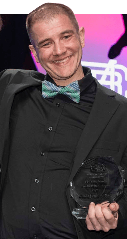 Justin Caine, wearing a black suit and shirt with a blue-and-green plaid bow tie, smiles while holding a clear, faceted award as he receives the MCSB Disability-Owned Small Business Award.