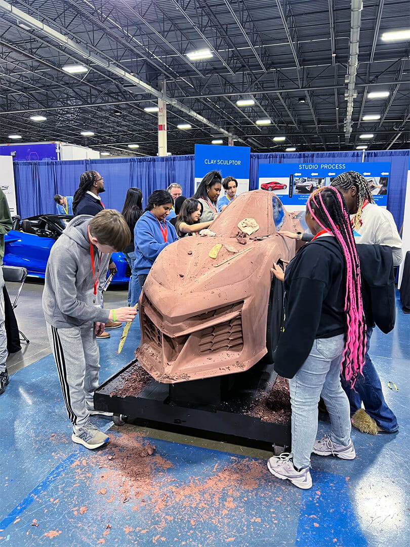 A group of participants at a MiCareerQuest event works together on a life‑size clay model of a car, using sculpting tools as clay pieces fall to the floor. Behind them are blue display panels labeled “Clay Sculptor” and “Studio Process” inside a large convention hall.