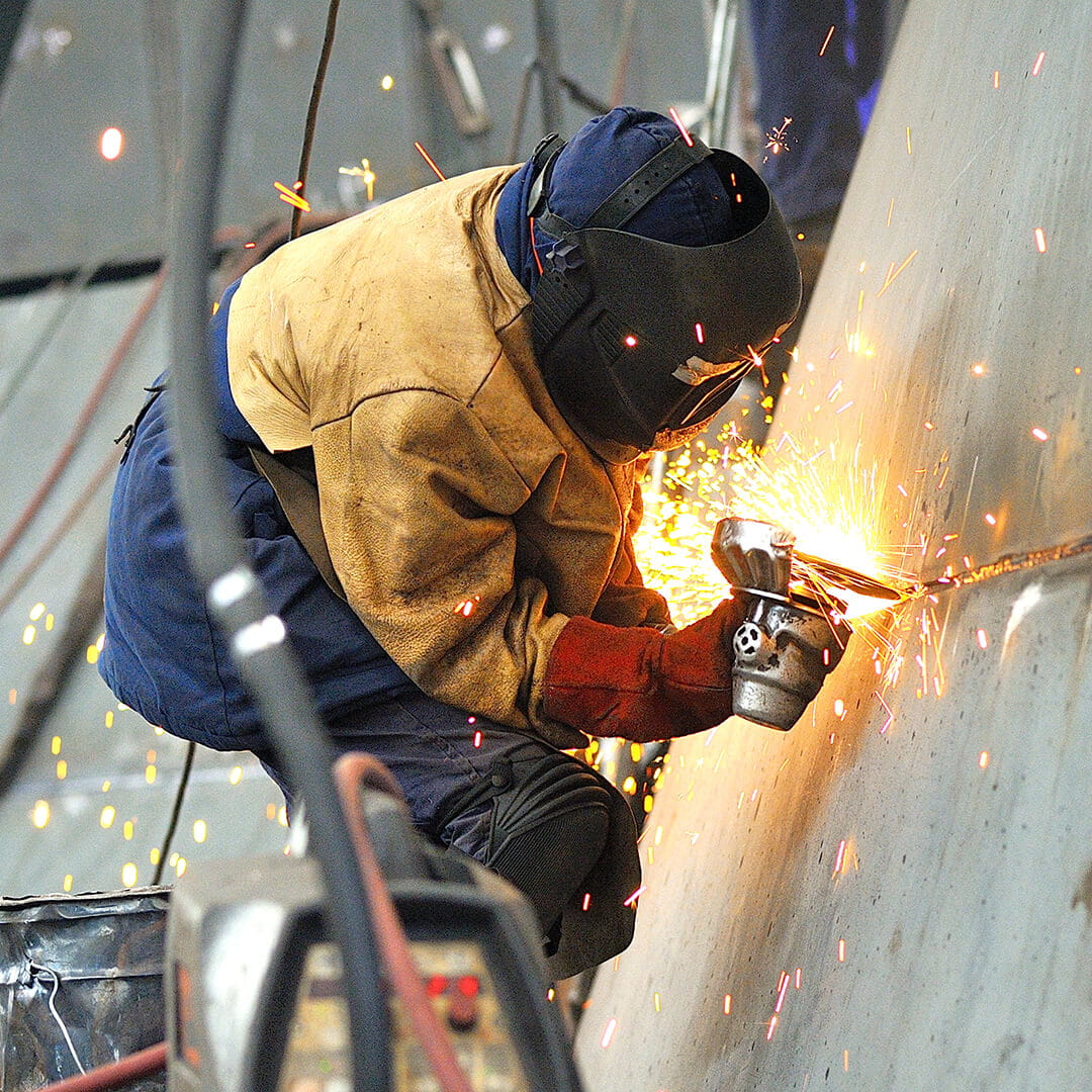 Person in protective gear using a power grinder on a metal surface, creating bright sparks in an industrial setting.