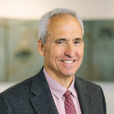 Joseph smiles wearing a light plum shirt and grey blazer