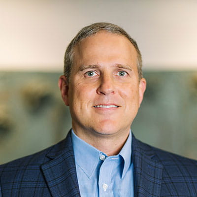 Jason smiles wearing a blue dress shirt and dark blue plaid blazer