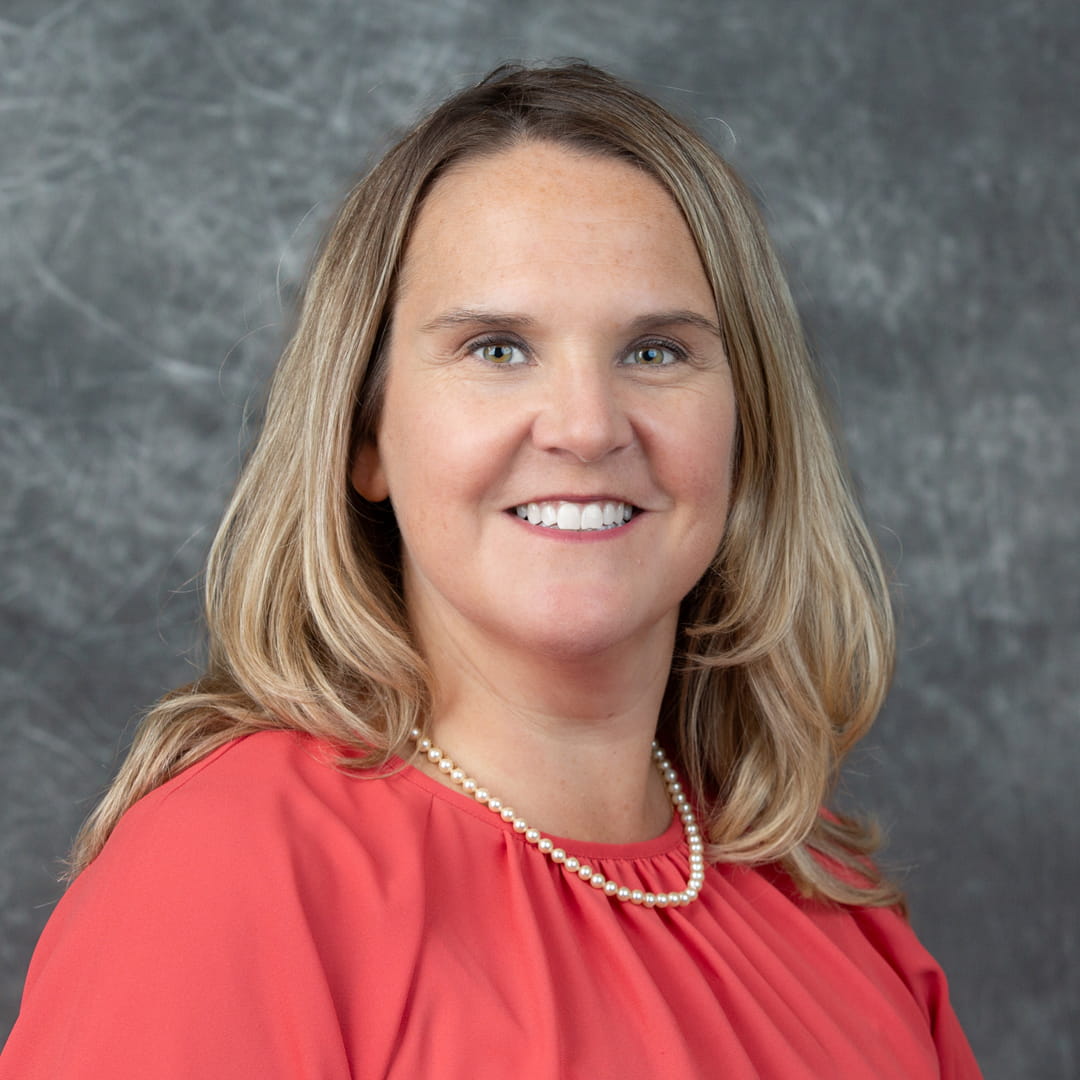 Stephanie Beckhorn, with shoulder‑length hair, shown in a professional head‑and‑shoulders portrait against a gray textured background. She is wearing a coral-colored top and a pearl necklace.