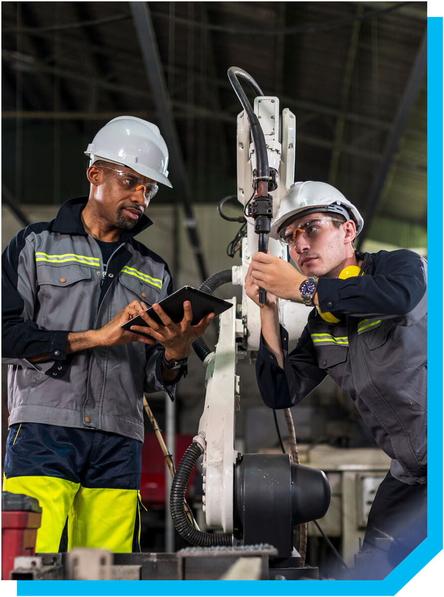 Two factory workers wearing hard hats, safety glasses, and protective jackets perform an inspection of robotic equipment in an industrial warehouse.