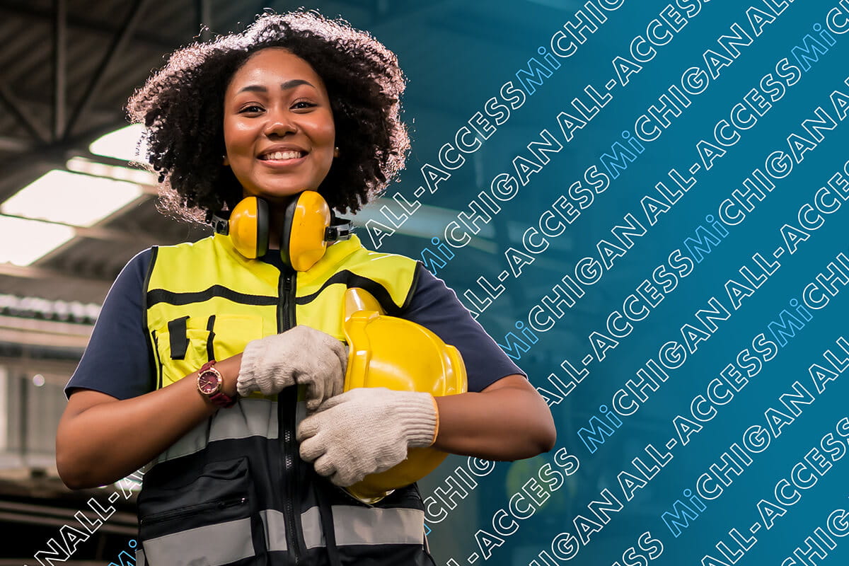 Smiling factory worker wearing a bright yellow safety vest, ear protection headphones around her neck, and gloves while holding a yellow hard hat in an industrial factory setting. The words "ALL-ACCESS MICHIGAN" are tiled diagonally in the background.