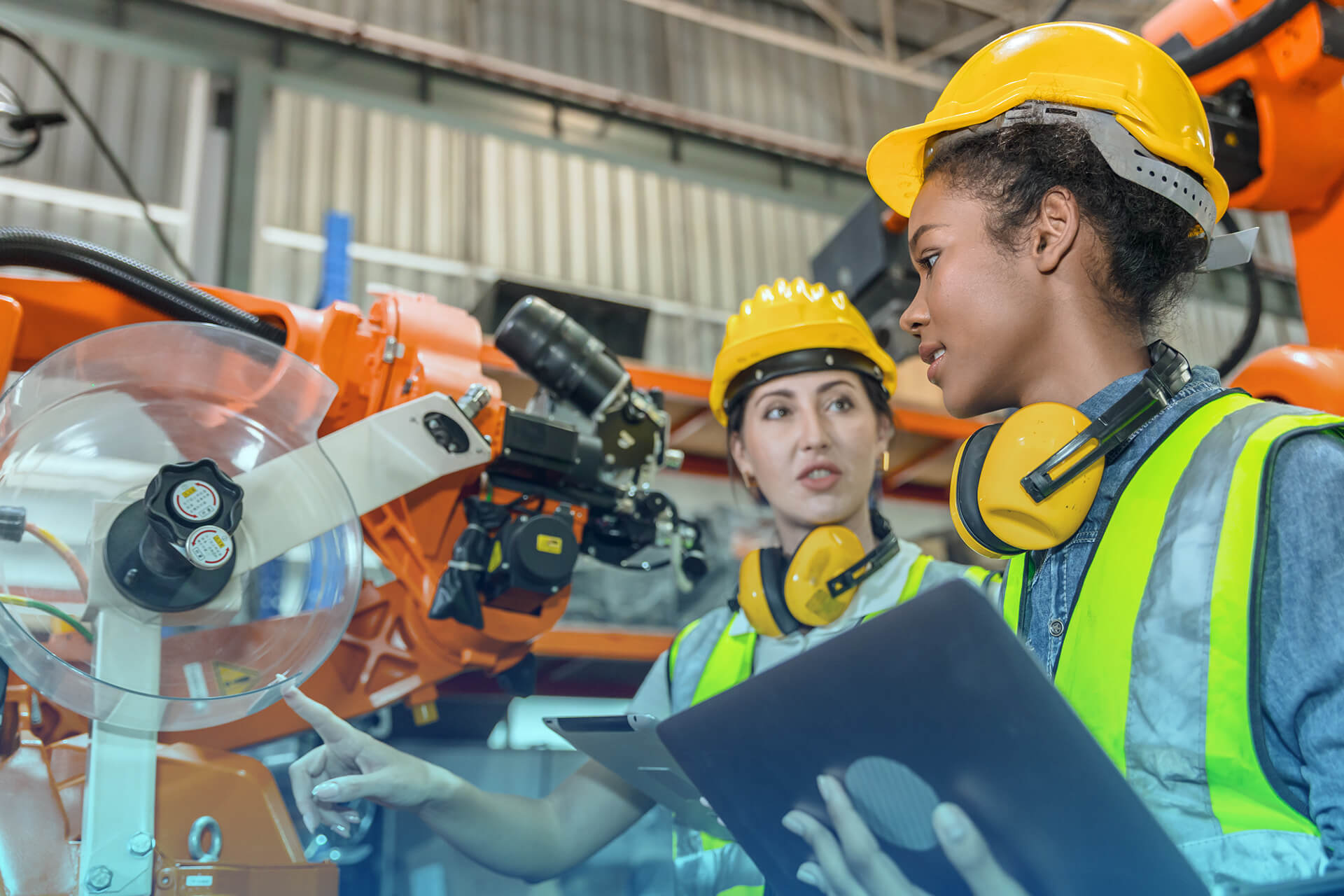 Two young engineers wearing yellow hard hats, safety vests, and protective headphones stand in front of an orange industrial robotic arm. One holds a laptop while the other points at the machine, appearing to discuss its operation in a factory setting.