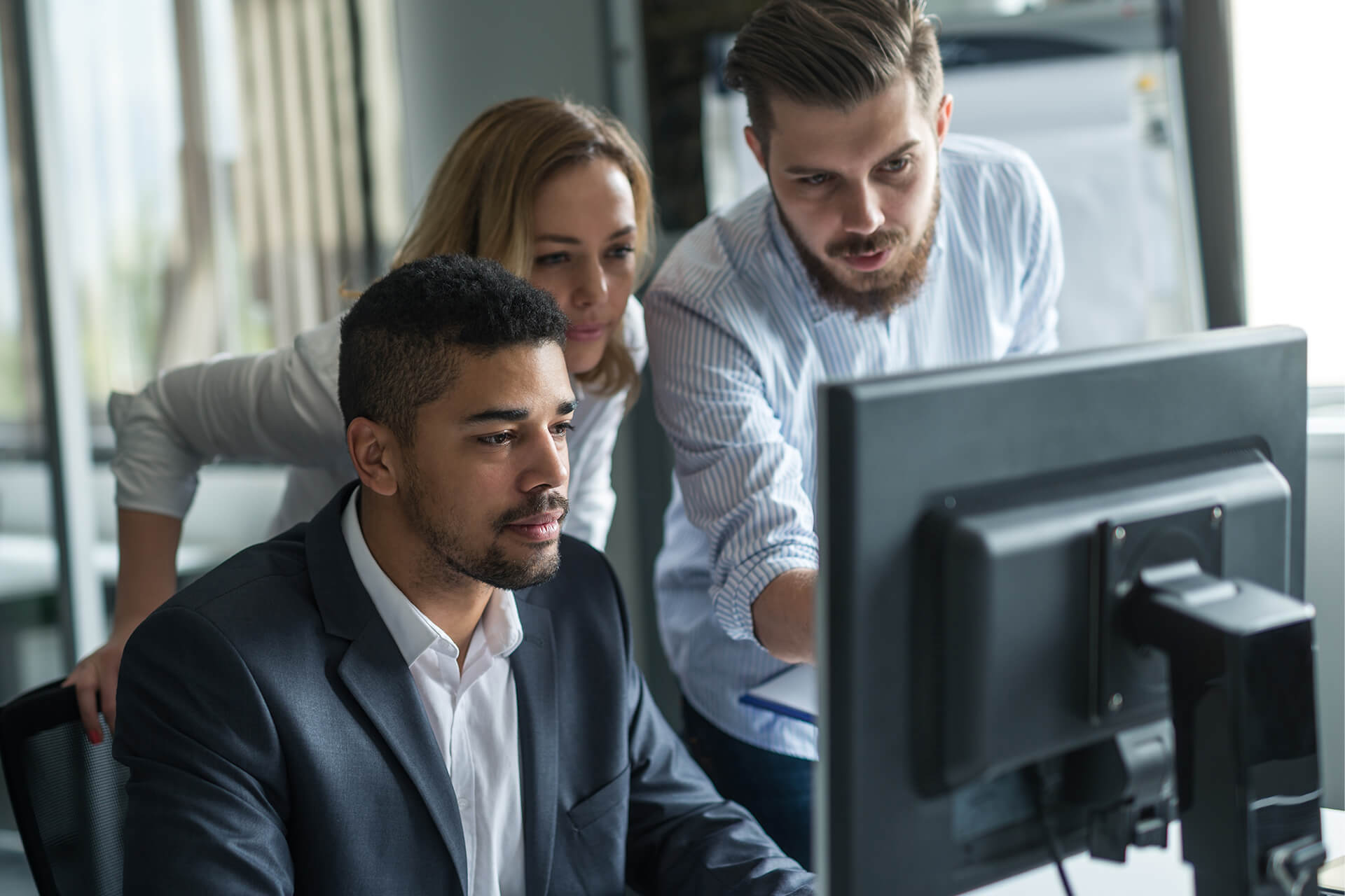 Three colleagues gathered around a computer monitor in a modern office. A man in a suit is seated at the desk, while a woman and another man lean in closely, looking at the screen with focus and collaboration.