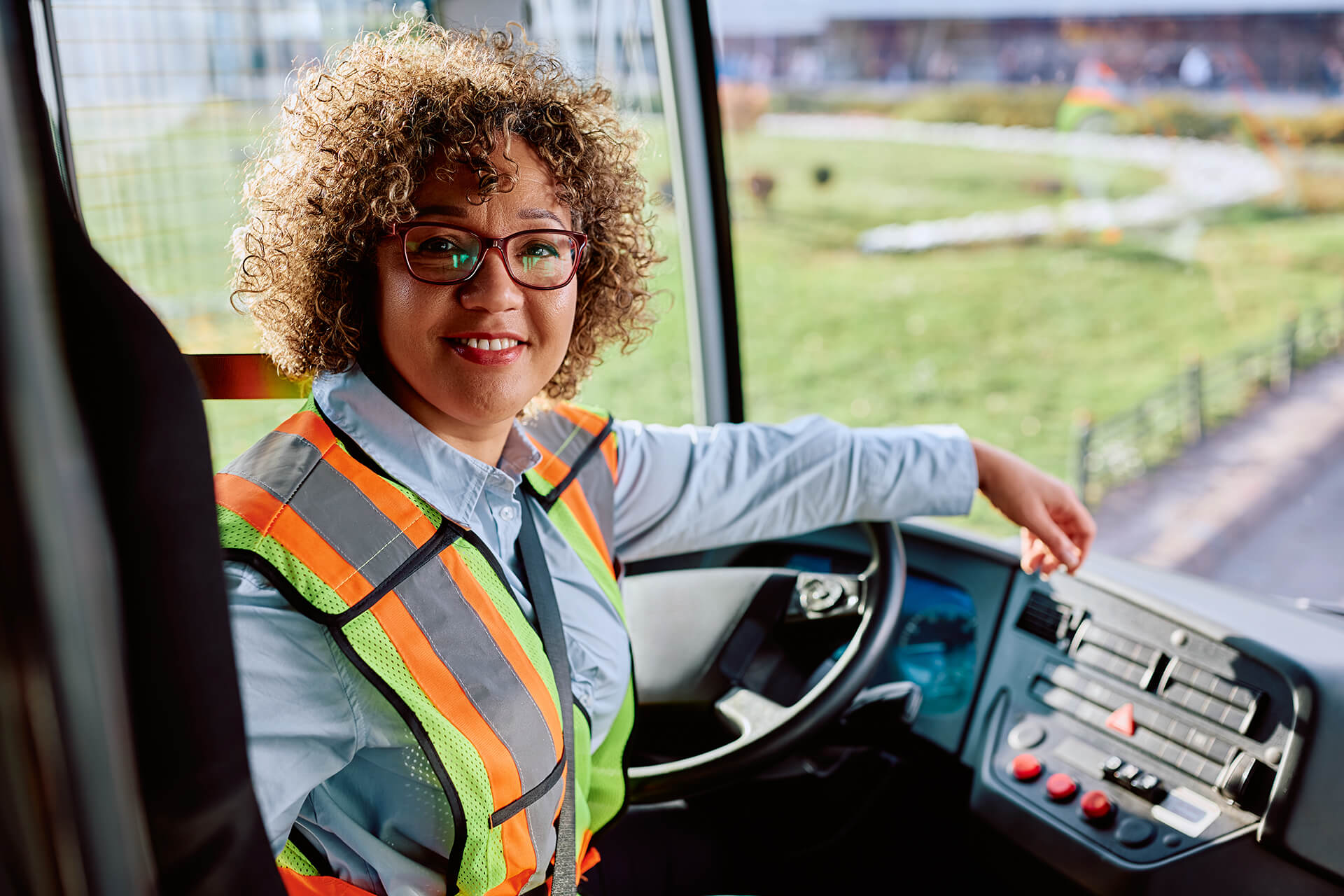 Bus driver smiling at the camera while seated at the driver’s seat, wearing a reflective safety vest and glasses, with one hand resting on the dashboard controls.