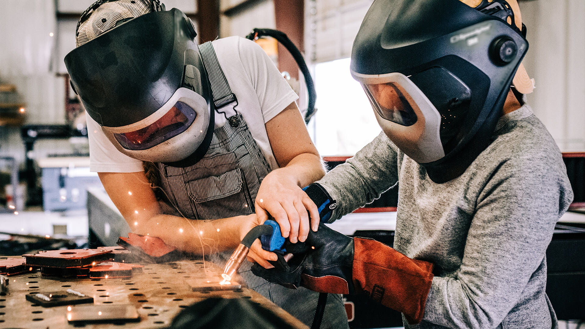 wo individuals wearing protective welding helmets and gloves work together in an industrial workshop while using a welding torch that emits bright sparks on a metal surface.