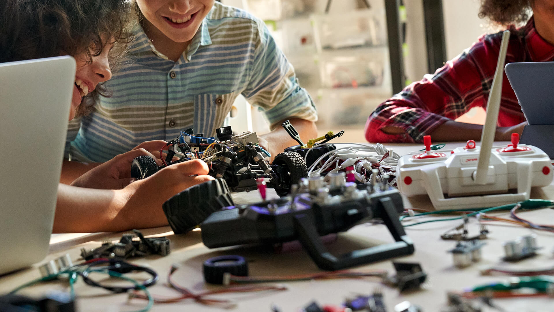 A group of young students sit at a table working on a small robotic vehicle, surrounded by laptops, remote controllers, wires, and electronic components. The scene shows hands adjusting parts of the robot and various tools spread across the workspace, highlighting a collaborative STEM or robotics activity.