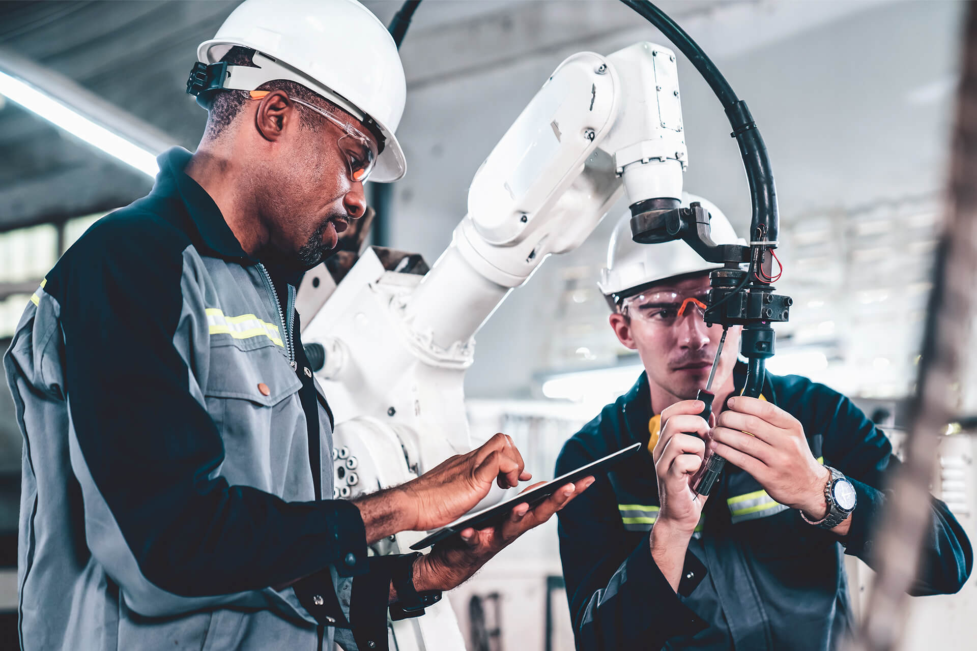 Two workers wearing protective helmets and safety glasses are operating and inspecting an industrial robotic arm. One worker is using a tablet, while the other adjusts wiring on the robotic machinery in a factory setting.