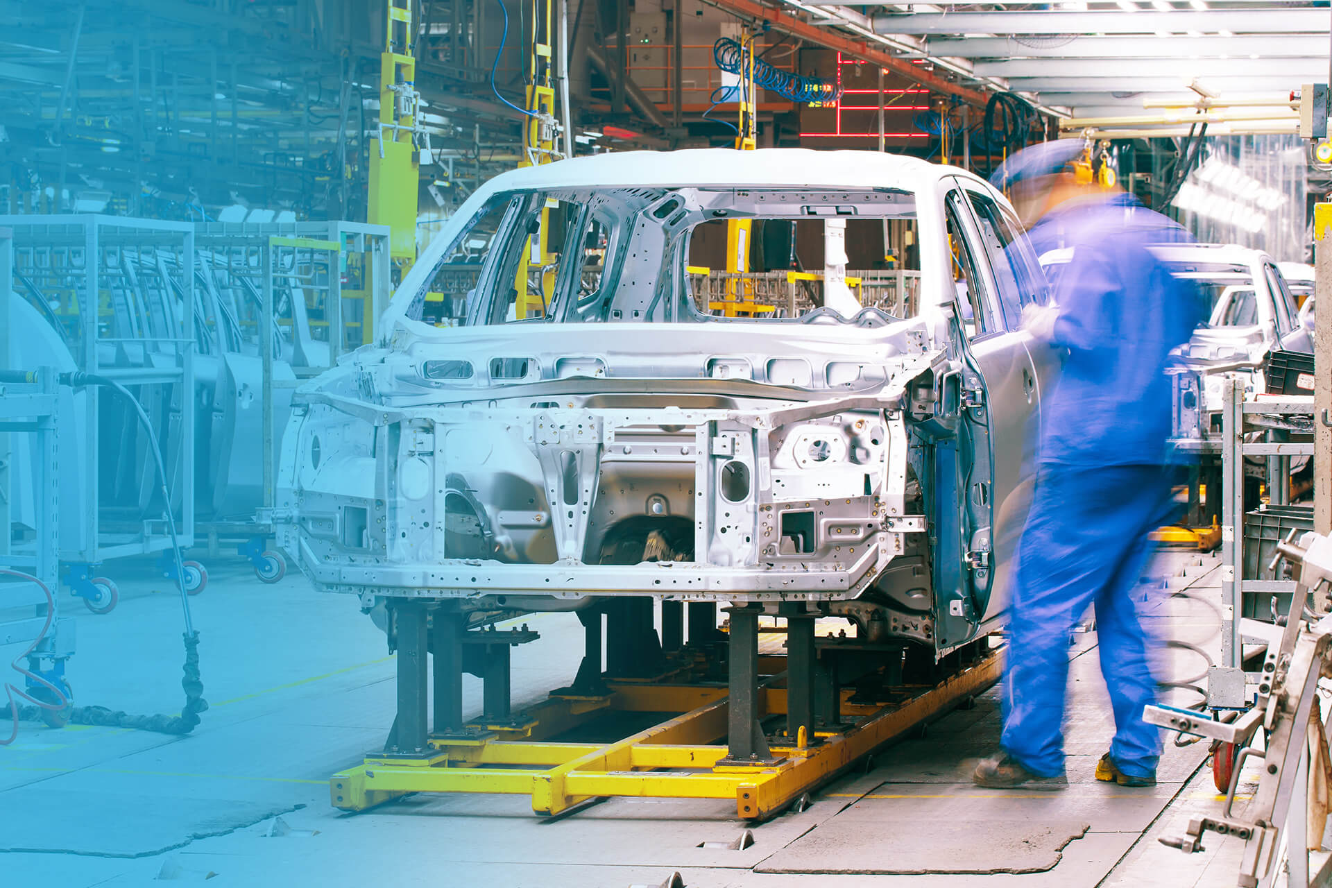 An automotive assembly line with partially built car bodies mounted on tracks. A worker in blue coveralls moves alongside the vehicles as they progress through the production process in the brightly lit factory.