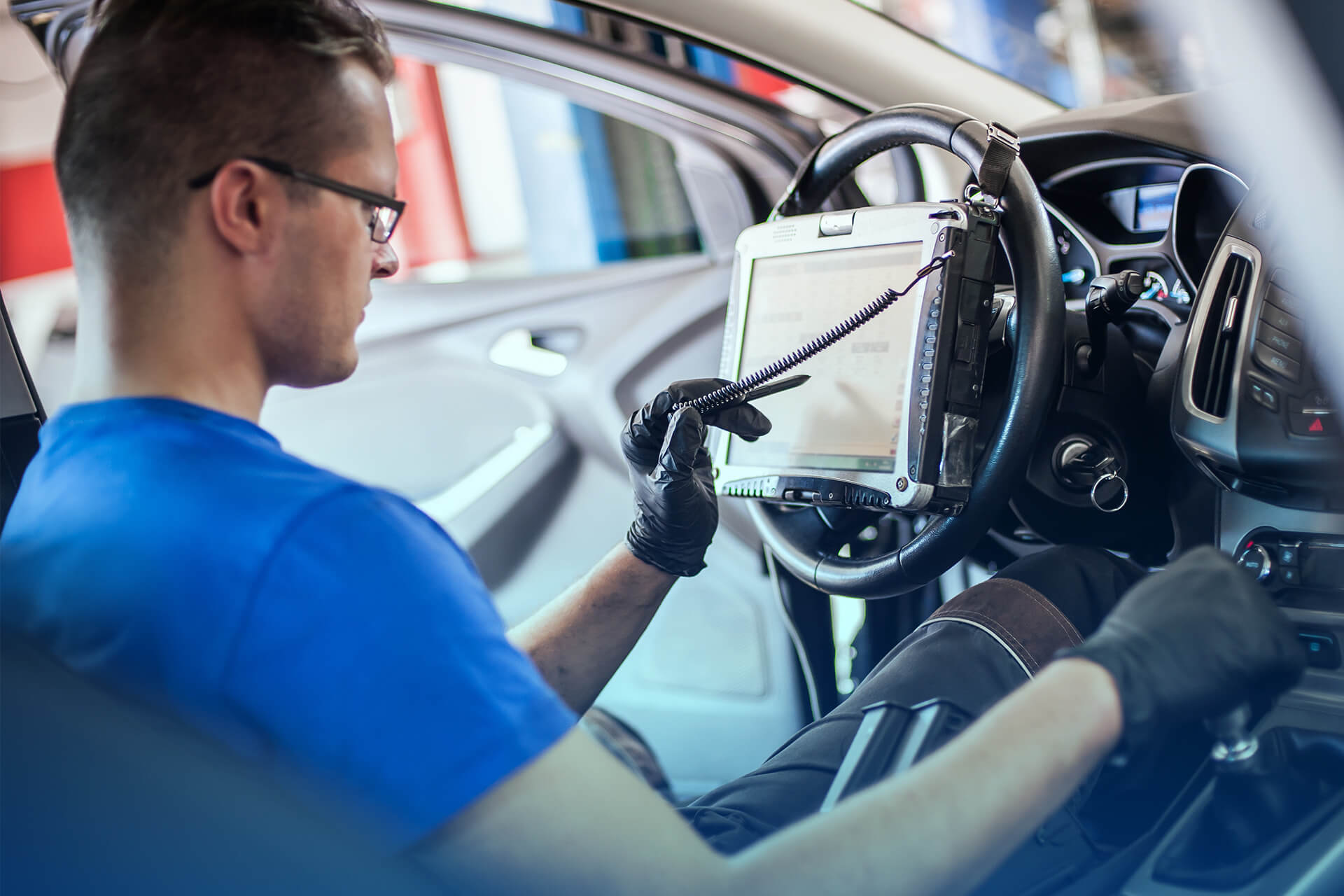 Automotive technician sitting inside a car using a diagnostic tablet connected to the vehicle’s onboard system, wearing black gloves and a blue shirt.