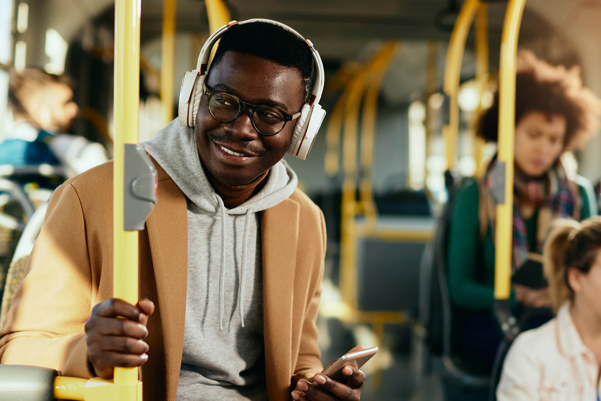 A man sitting on a city bus holding a smartphone and smiling while wearing headphones and glasses. He is dressed in a tan coat over a gray hoodie, with other passengers visible in the background.