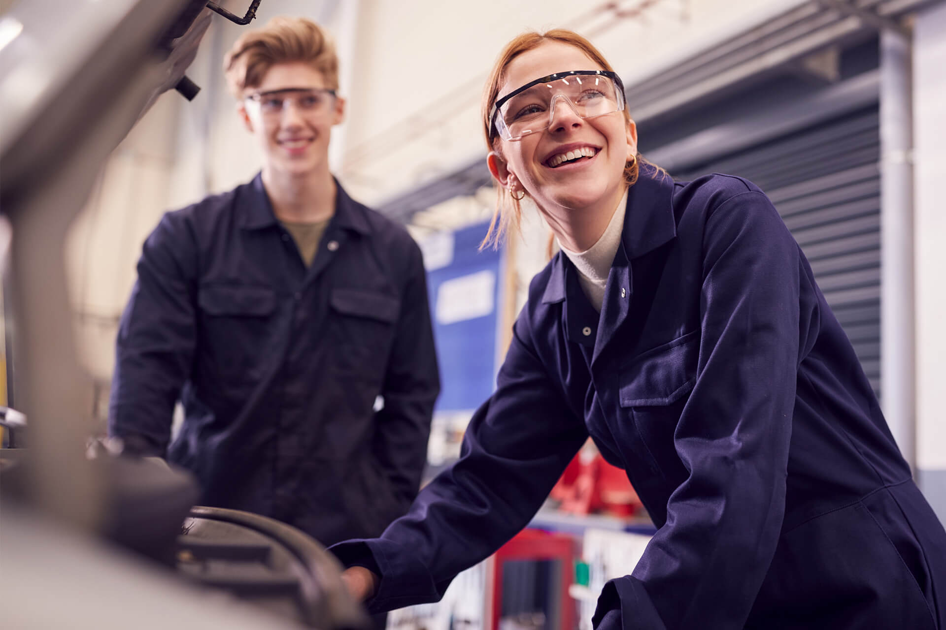 Two young people in navy blue coveralls and safety glasses standing by machinery in a workshop. The woman in the foreground is smiling brightly while working on equipment, with her colleague watching in the background.