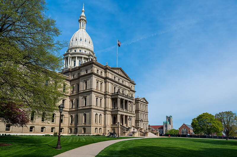 The Michigan State Capitol building stands under a clear blue sky, with its white dome and flagpole centered above the historic structure. A curved pathway leads through the green lawn toward the entrance, with trees framing the scene.
