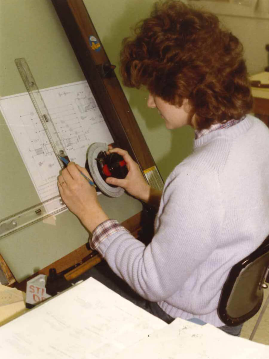 Woman seated at a drafting table, using a protractor and ruler to create precise lines on a technical blueprint.