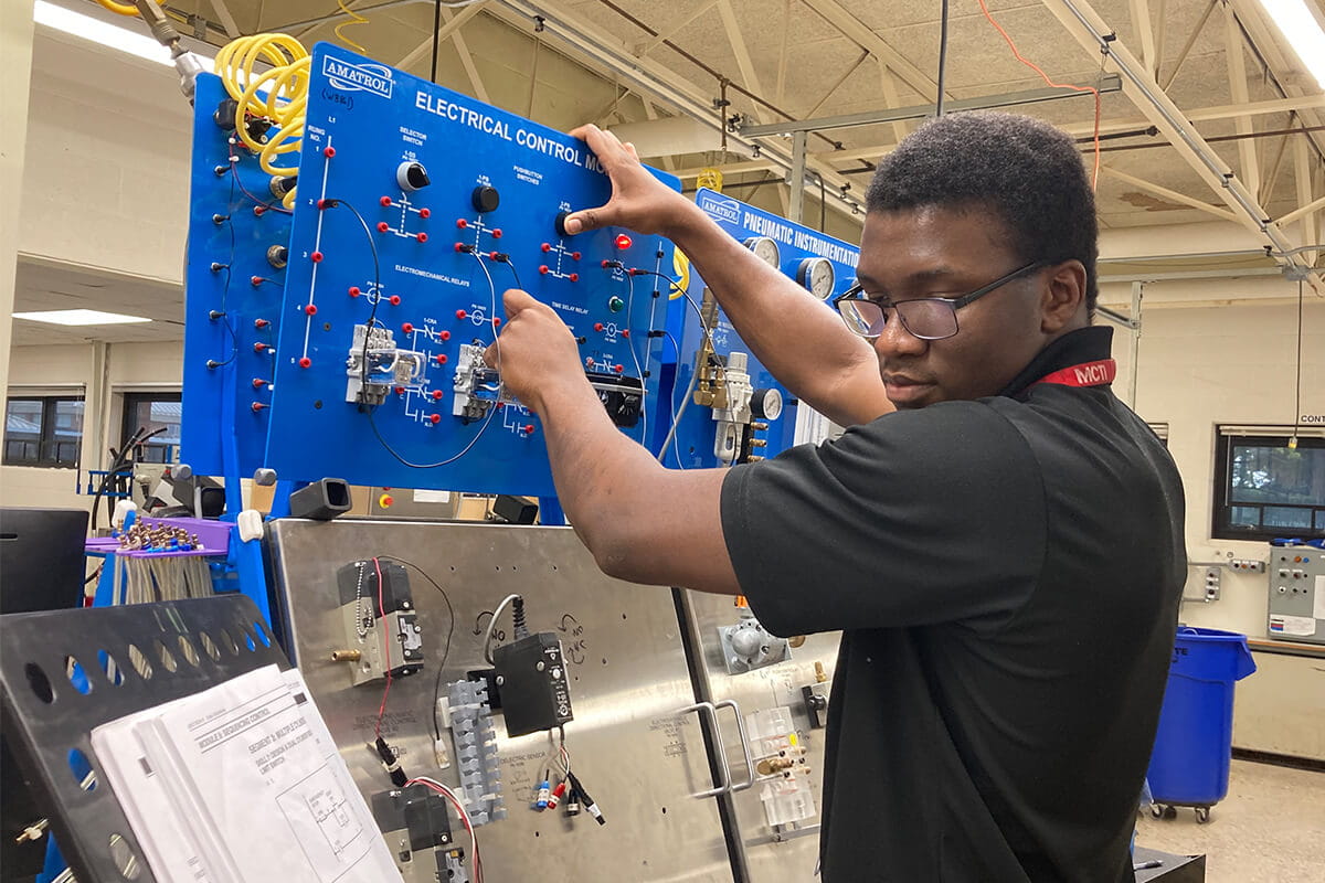 A modern-day MCTI student working on an electrical control module, connecting wires on a blue training panel in a technical lab setting.