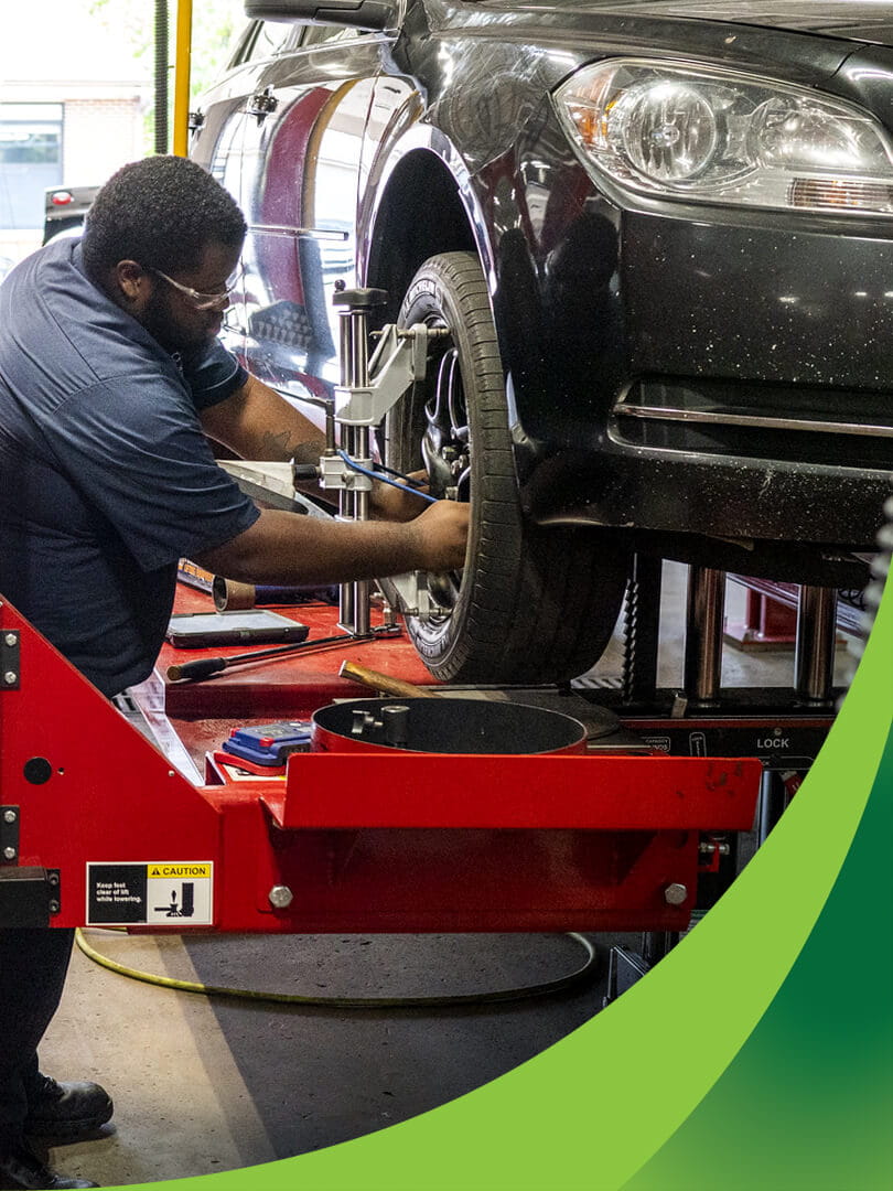 MCTI Automotive Technology student inspecting the wheel assembly of a car that is elevated on a hydraulic lift in an auto repair shop.