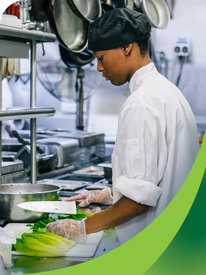 MCTI Culinary student dressed in a chef's coat and cap using a classic chef's knife to prepare lettuce on a cutting board in a commercial kitchen.