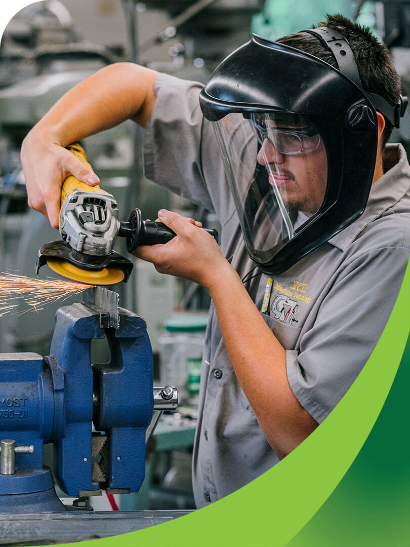 MCTI Machine Technology student wearing a protective face shield while using a handheld grinding wheel to smooth the surface of a piece of scrap metal in a fabrication shop.