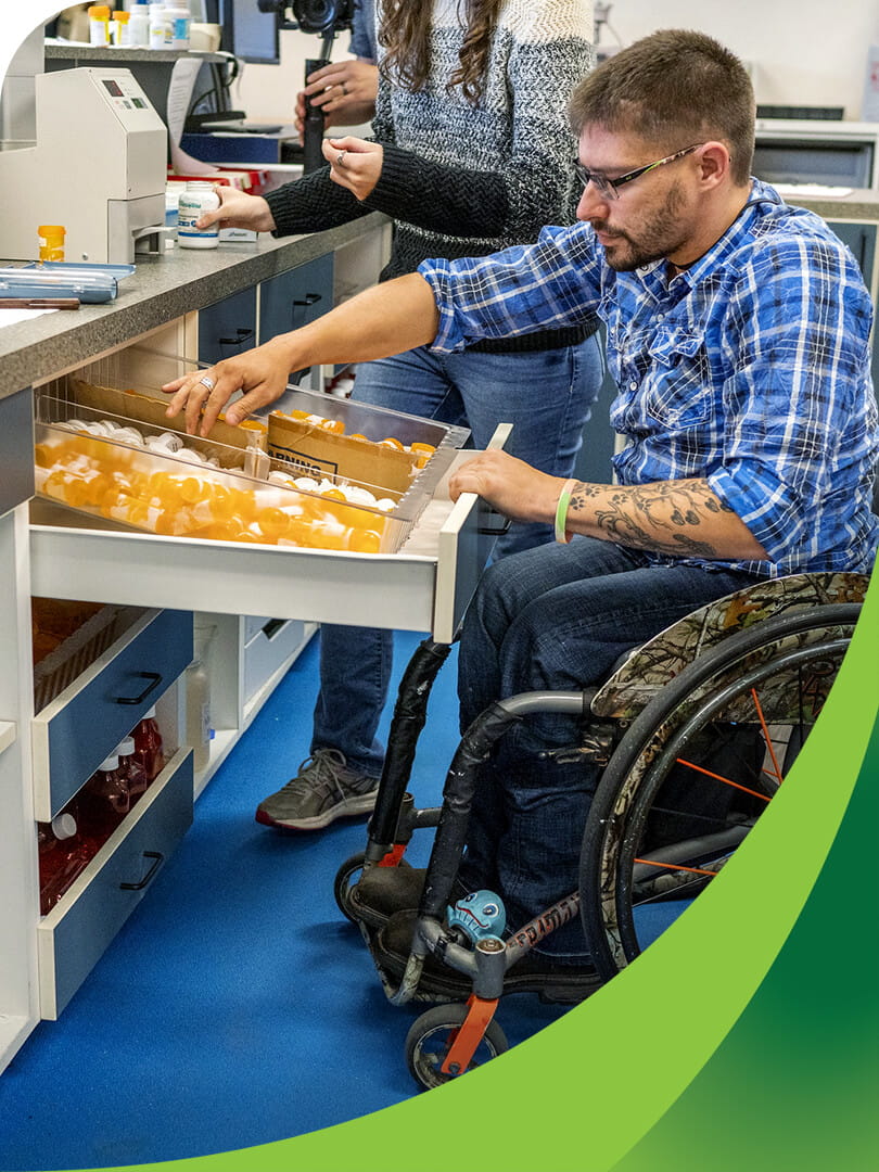 MCTI Pharmacy Technician student in a wheelchair opening a drawer of medication bottles beneath a pharmacy counter in preparation for dispensing medications.