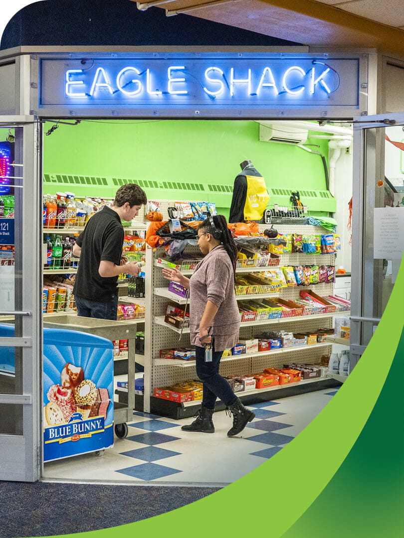 MCTI Retail Marketing students working in the Eagle Shack, MCTI's on-campus convenience store, with a brightly lit neon blue sign displaying the store's name above the entrance.