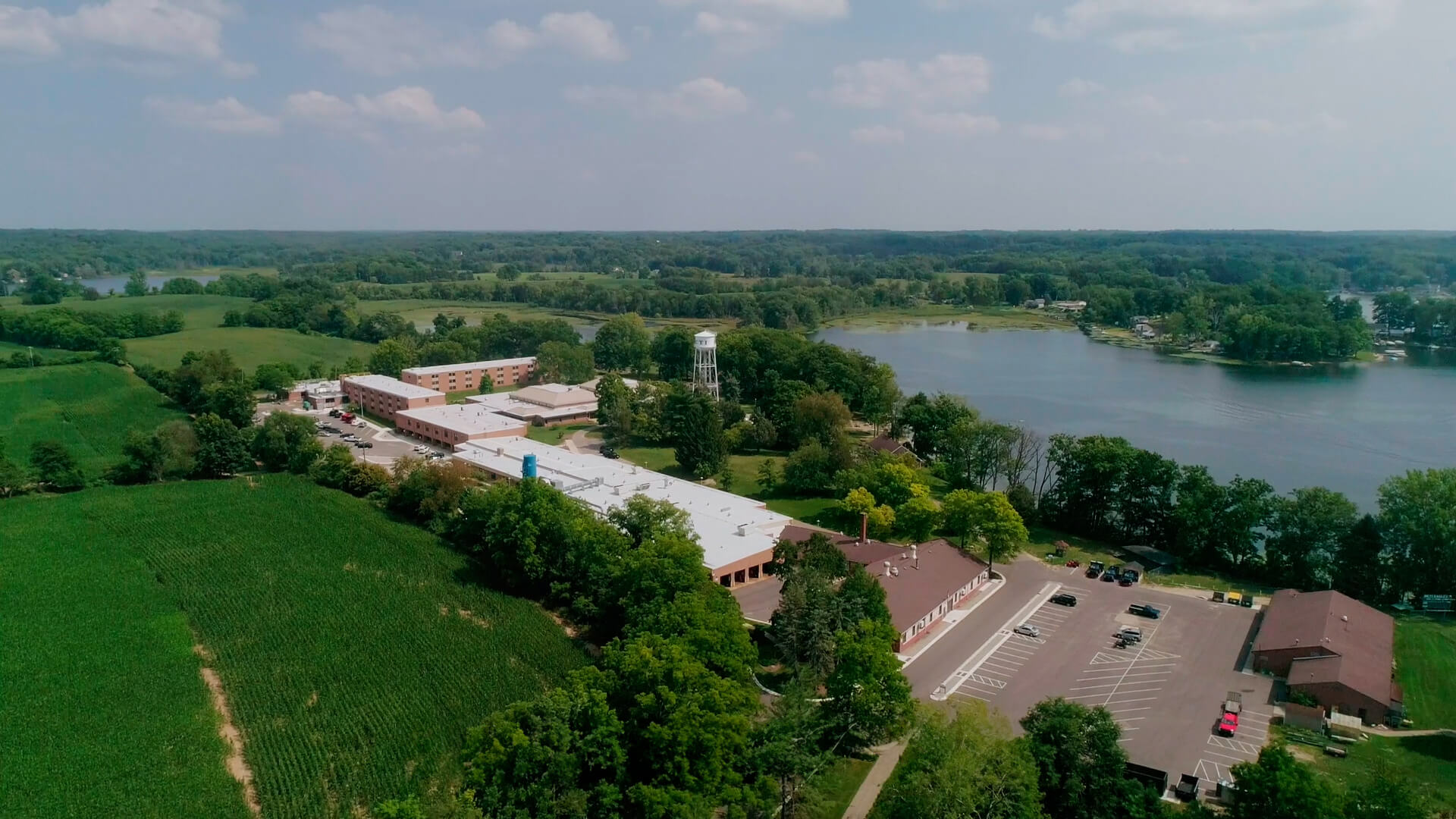 An aerial view of MCTI's main campus in Plainwell, Michigan. Surrounded by lush green fields, trees, and Pine Lake, the campus features multiple buildings, parking lots, a water tower, and a serene landscape of farmland and wooded areas.