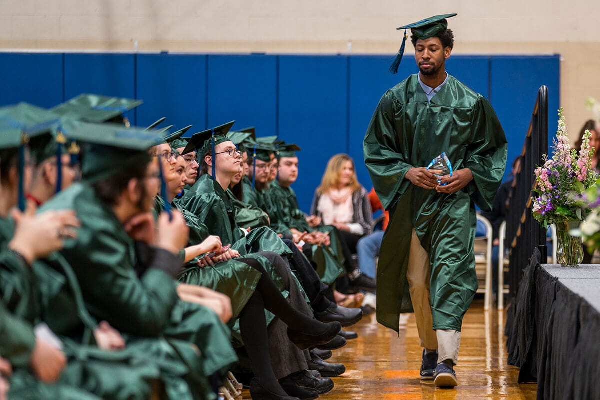 An MCTI graduate in a cap and gown walks confidently in front of the stage holding a glass award plaque, with seated peers from the graduating class looking on.