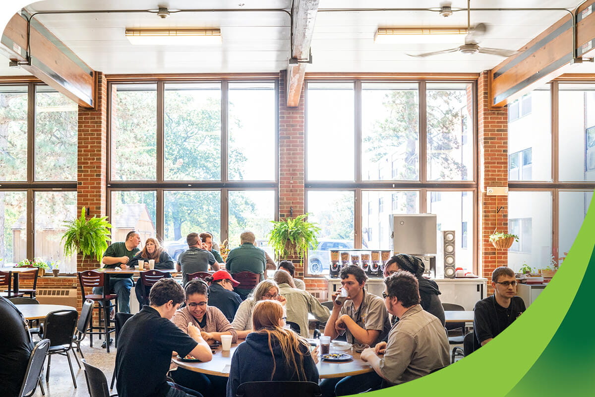 Students engaging in conversation around a table in a brightly-lit cafeteria with floor-to-ceiling windows.