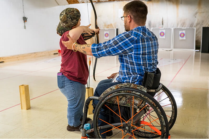 An MCTI student in a wheelchair assists a fellow MCTI student, a little person, in adjusting their archery form as both students practice together in an inclusive environment.