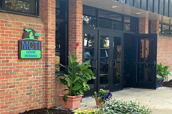 Entrance to the MCTI building with a sign reading 'MCTI Leisure Entrance' mounted on a brick wall. The entrance features glass doors, and there are potted plants with green foliage and flowers near the doorway.