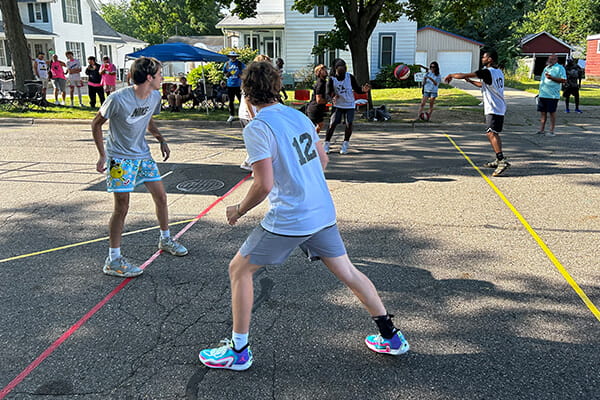 Group of people playing a basketball game on a residential street, with two players in the foreground actively defending. Spectators and other participants are visible in the background, along with houses and trees on a sunny day.