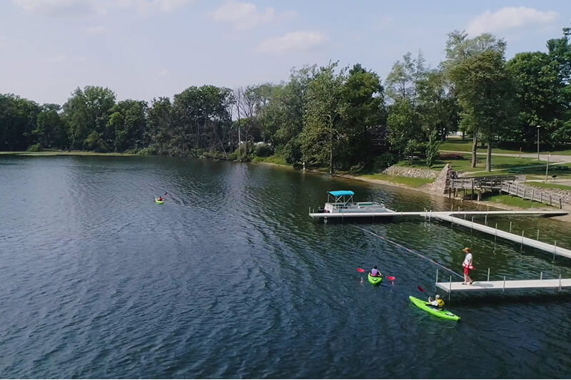 Aerial view of MCTI students kayaking near the shoreline docks of Pine Lake on a sunny day, with calm waters and surrounding natural scenery.