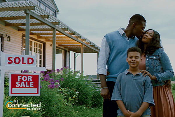 man and woman kissing with school aged child in front of them all standing next to a house with a sold sign