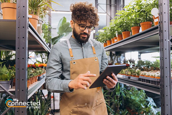 person holding a tablet in a greenhouse next to shelves of plants
