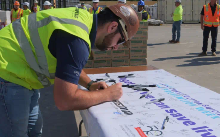 Construction worker in safety gear signs a banner that reads “Together, we can save lives” during a workplace safety event.