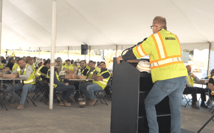 MIOSHA representative addresses a group of construction workers seated under a tent at a safety event.