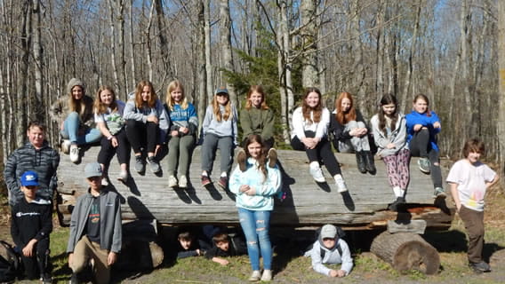 a group of students sitting outside on a large log as some stand around it