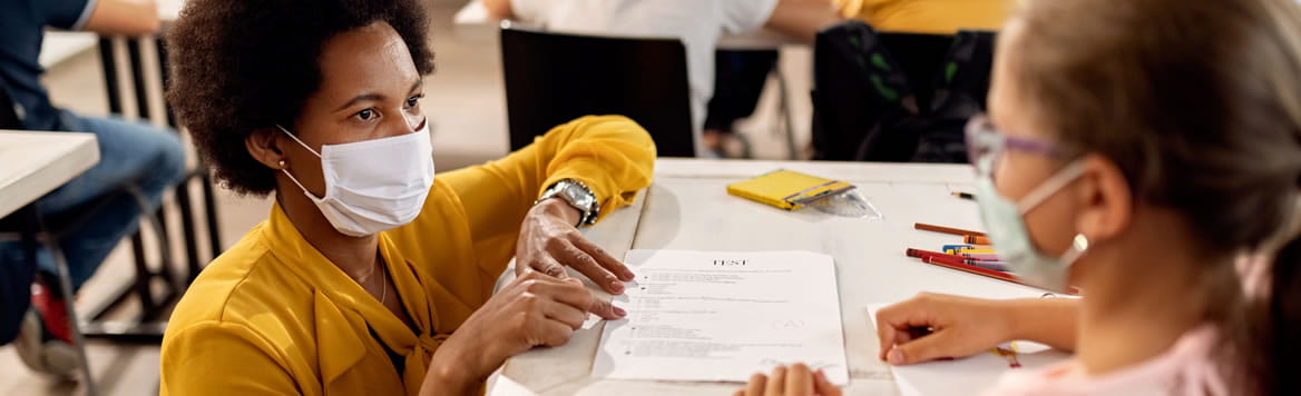 teacher in a classroom kneeling at a young student's desk going over a test with student