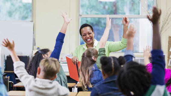 teacher in classroom with young students all raising a hand