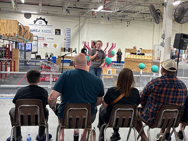 A robotics student speaks to an audience at MiSTEM's FIRST Robotics grand opening with the robotics setups featured behind him