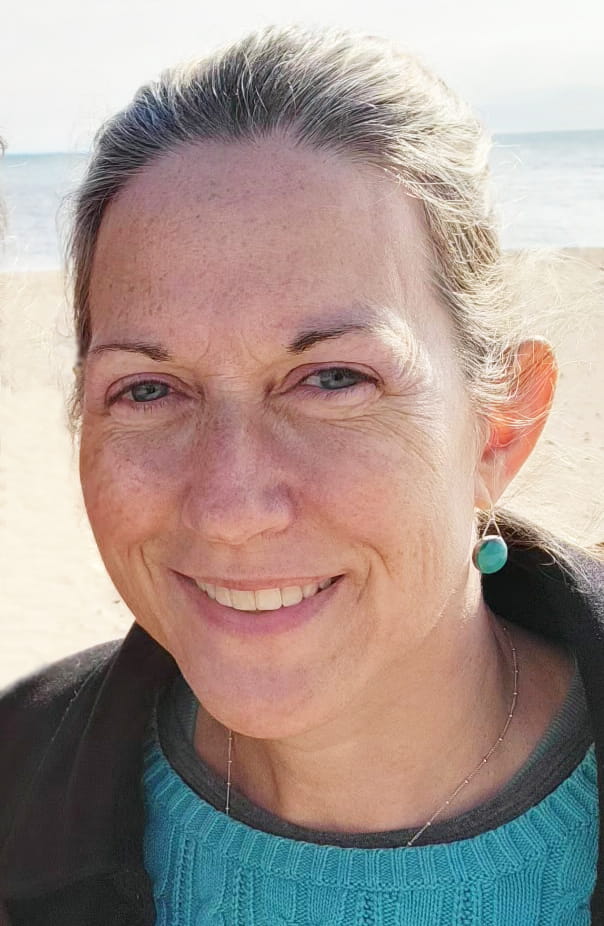 Portrait of Heather smiling on a brightly-lit day with a beach and water in the background. She is wearing a turquoise sweater and black fleece jacket with turquoise earrings.