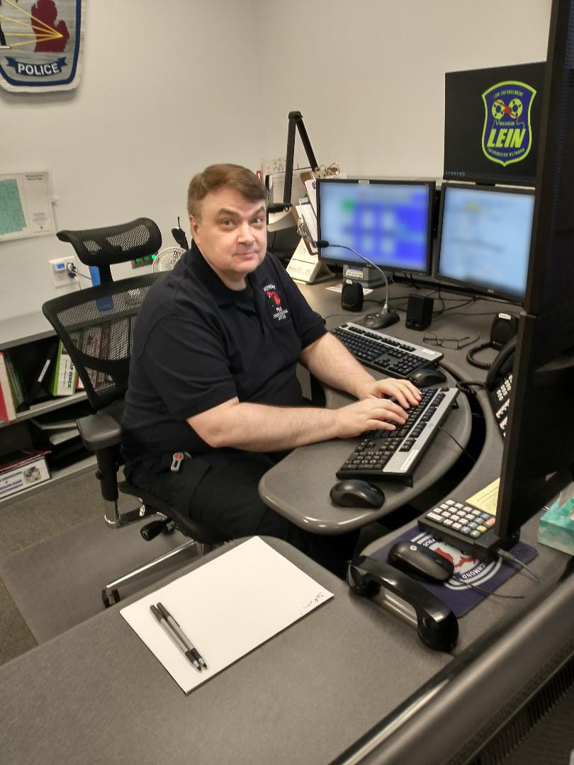 Earl Baranowski dressed in a black polo shirt, seated in an office chair and partially surrounded by computer monitors as he prepares to type at a 911 dispatch workstation.
