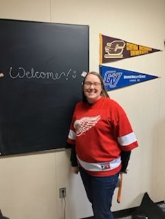 Dena, wearing a red Detroit Red Wings hockey jersey, stands beside a chalkboard with the word “Welcome!” written on it in white chalk. Two college pennants — one for Central Michigan University and one for Grand Valley State University — hang on the wall behind her.