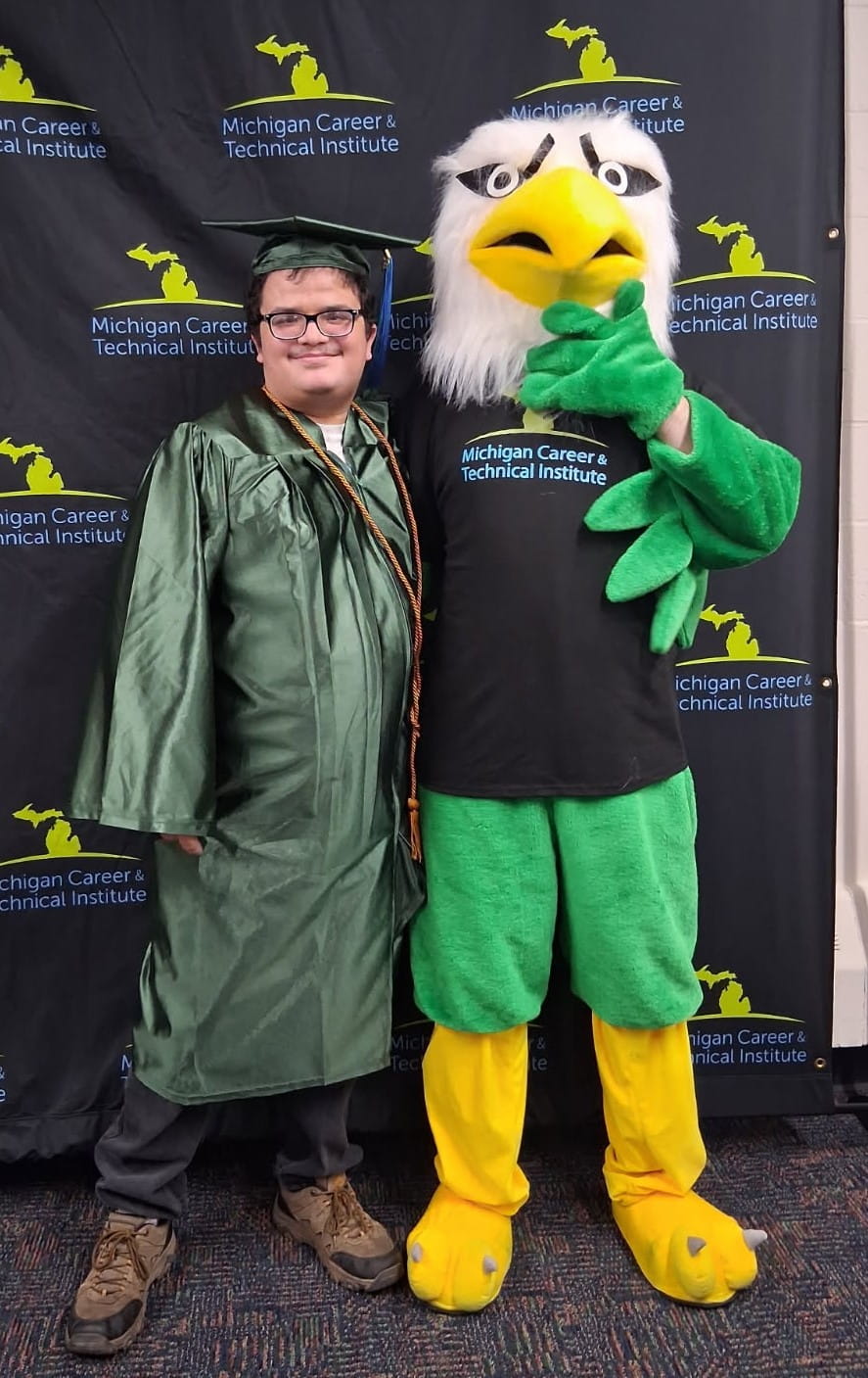 Clayson, wearing a green graduation cap and gown with honor cords, smiles as he poses beside a person in a green and yellow eagle mascot costume. The mascot has a large yellow beak, white feathers, and is wearing a black Michigan Career & Technical Institute T-shirt. They are standing in front of a step-and-repeat backdrop with the Michigan Career & Technical Institute logo.