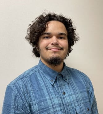 Cris, a customer of Michigan Rehabilitation Services and graduate of Michigan Career & Technical Institute (MCTI), standing in front of a plain light-colored wall and smiling at the camera. Cris has short curly hair, a trimmed beard, and is wearing a blue plaid button-up shirt.