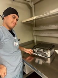 Bryan wearing scrubs and standing in front of sterilization machine. 